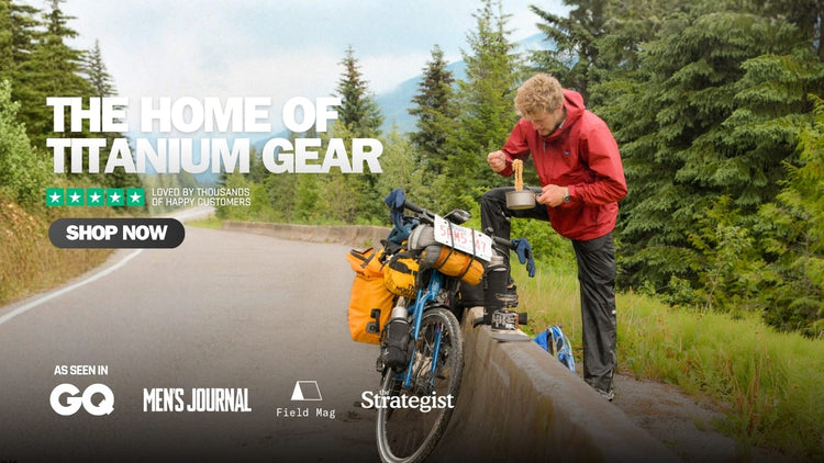 A man in a red jacket stands beside a fully packed bicycle on a forest road, holding a container near the bike rack. Large text reads 'THE HOME OF TITANIUM GEAR' with a 'SHOP NOW' button, and logos for GQ, Men's Journal, Field Mag and Strategist appear along the bottom.