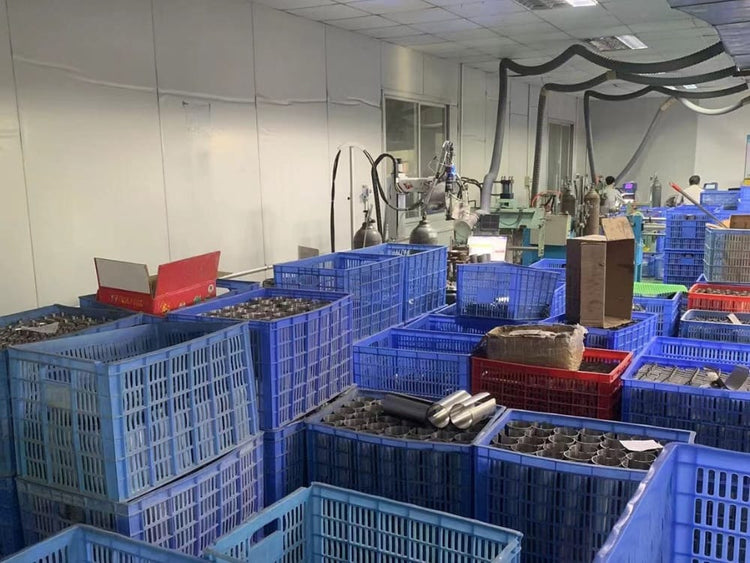 A factory workshop floor filled with blue plastic crates containing titanium gear, with industrial machinery and workers visible in the background.