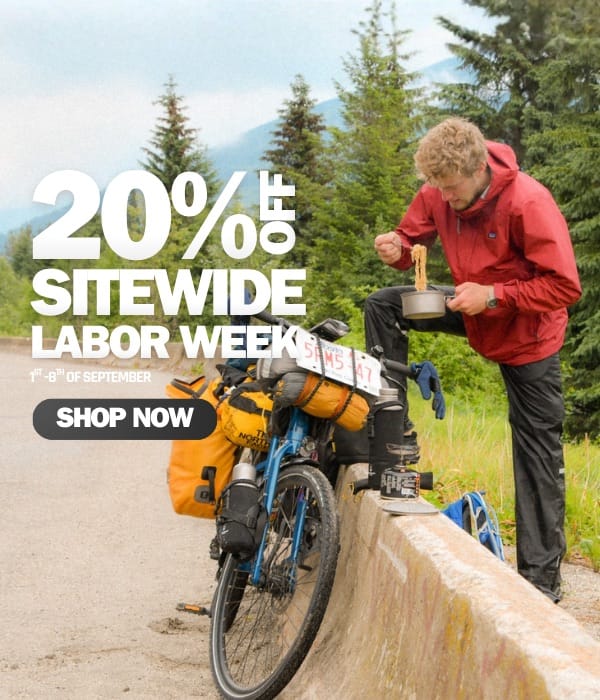 A man in a red hooded jacket and black pants stands beside a fully loaded touring bicycle on a road, eating from a bowl. Large promotional text reads '20% OFF SITEWIDE LABOR WEEK' with a 'SHOP NOW' button, set against a forest and mountain background.