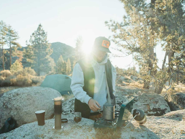 Person camping in a forest setting with cooking equipment making coffee