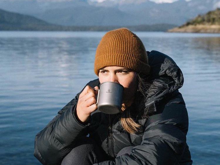 Person drinking from a mug by a lake with mountains in the background
