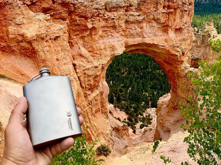 Hand holding a silverant flask in front of a natural rock arch with greenery