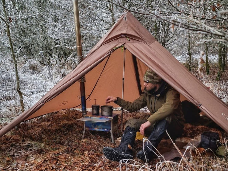 Person camping in a snowy forest with a tent and stove.