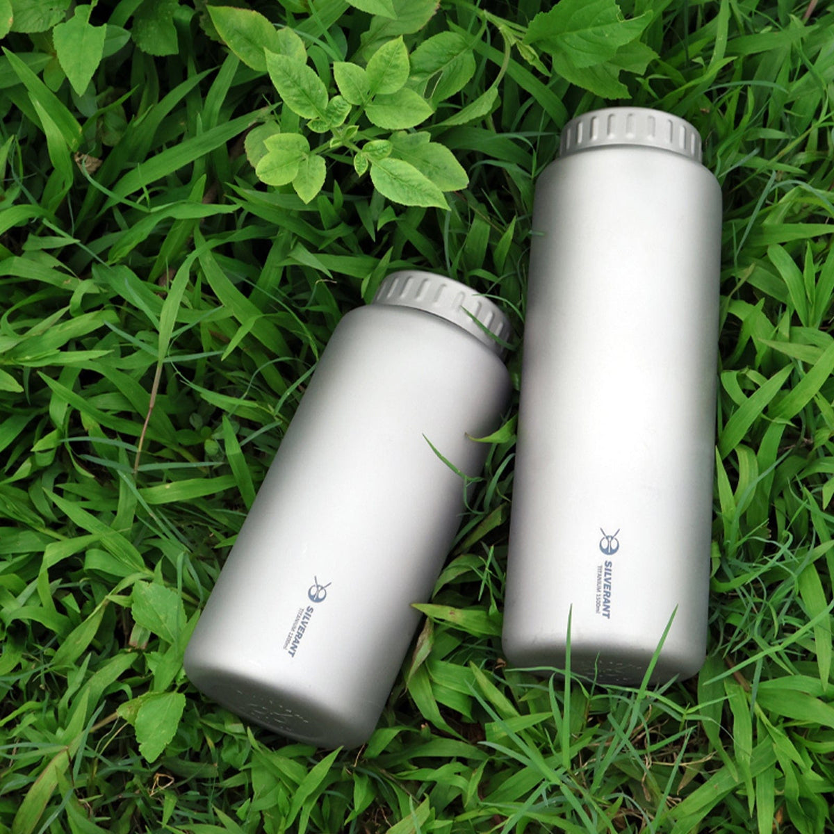 SilverAnt Ultralight Large Titanium Water Bottle (Wide Mouth) - In-Use (both sizes laid on the grassy ground, lifestyle photo, no thermal sleeve, silverant branding, titanium sandblasted finish)