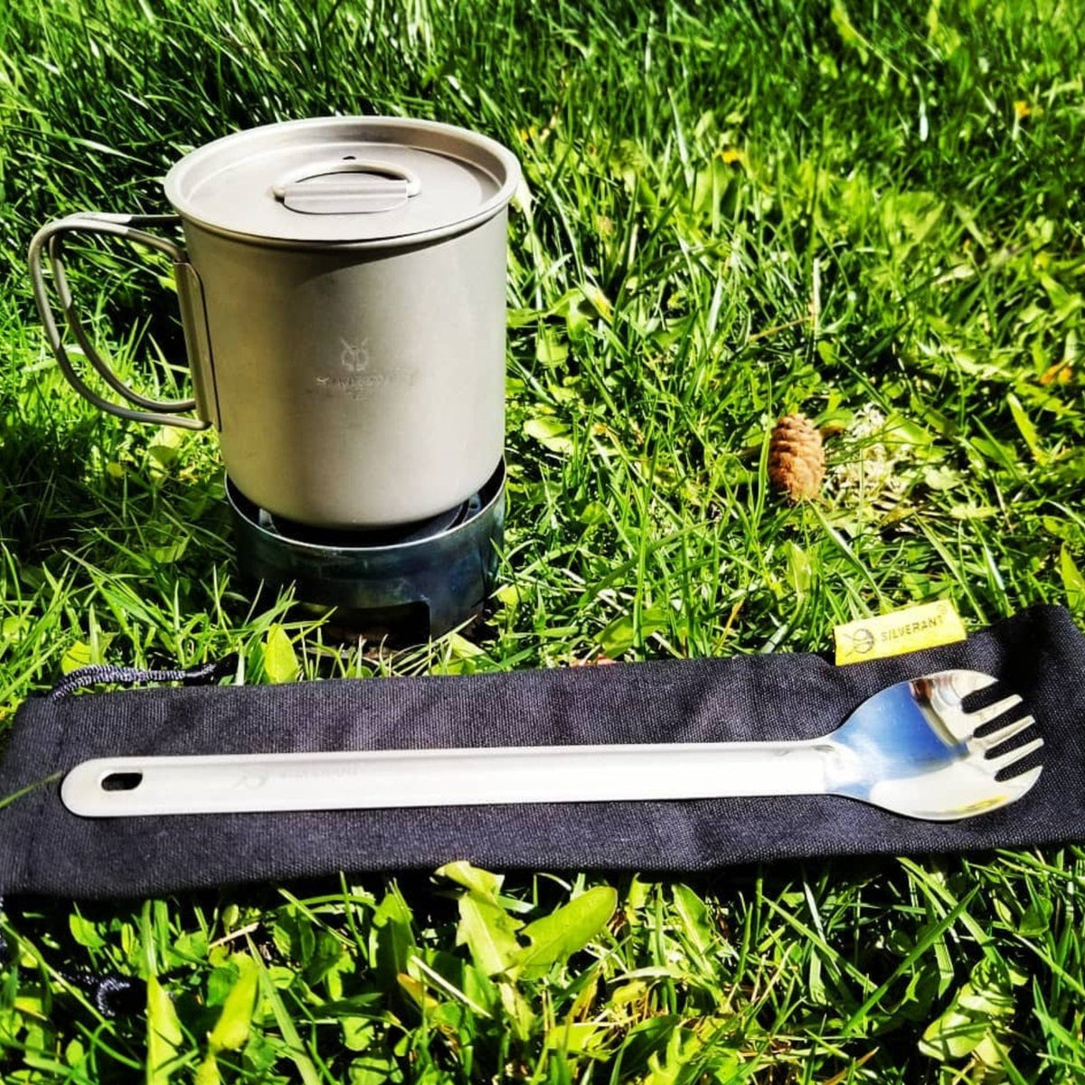 SilverAnt Ultralight Long Handle Titanium Spork Polished - actual product photo, real life use, lifestyle shot, in use, set on the grassy floor/ground with a titanium cup beside it