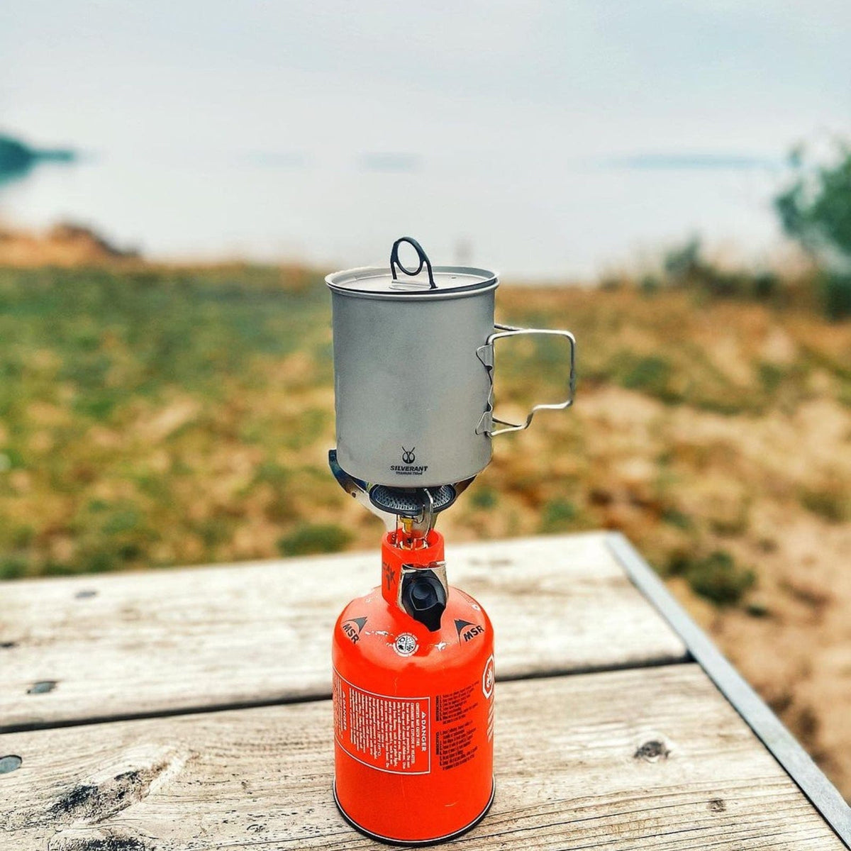 SilverAnt Ultralight Titanium Pot 750ml/25 fl oz with Lid - Actual product photo, real life still, lifestyle shot, placed on top of gas stove on a wooden table outdoors, camping scene