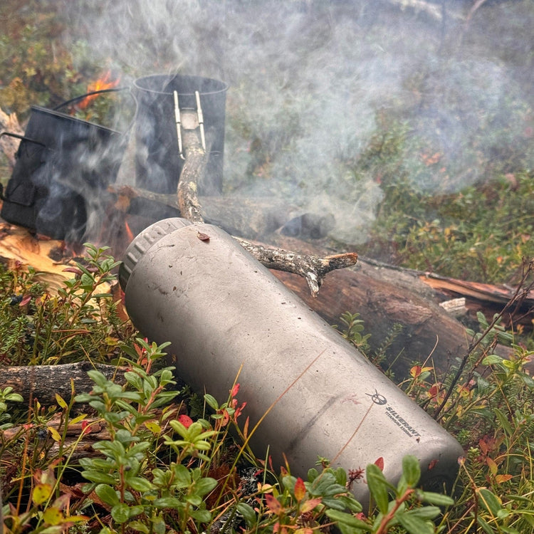 
                  
                    SilverAnt X 5Athlon Nomads Trails Collaboration - Large Titanium Wide Mouth Water Bottle laid down on grassy ground, smoke in the background from nearby campfire
                  
                